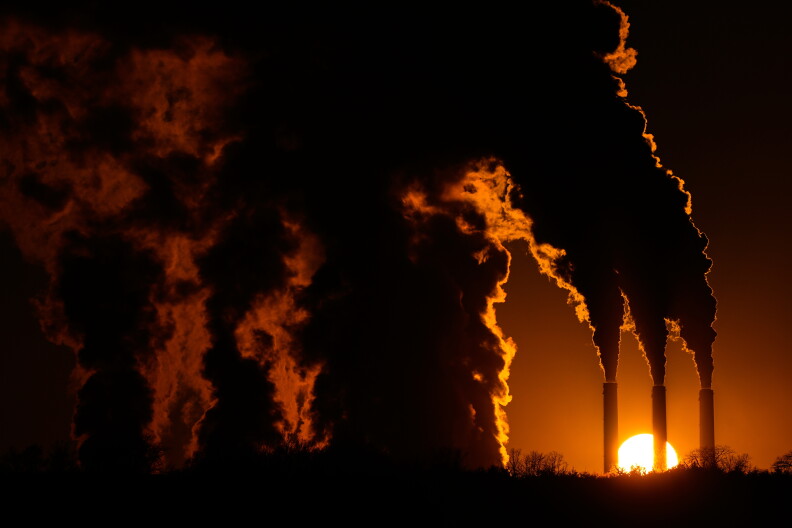 Several smoke stacks billow smoke as the sun sets behind them, shading them in an orange glow.